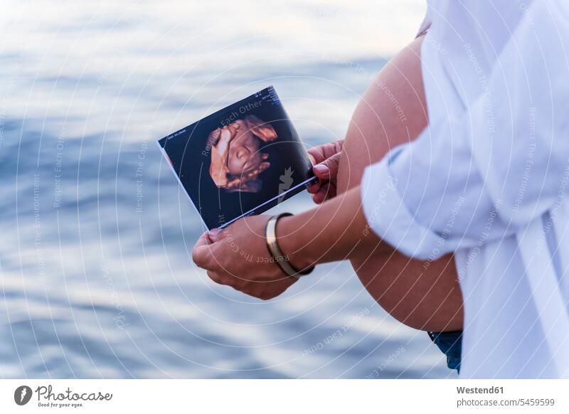Mittelsektion einer schwangeren Frau, die ein Sonogramm hält, während sie gegen das Meer steht Farbaufnahme Farbe Farbfoto Farbphoto Außenaufnahme außen draußen