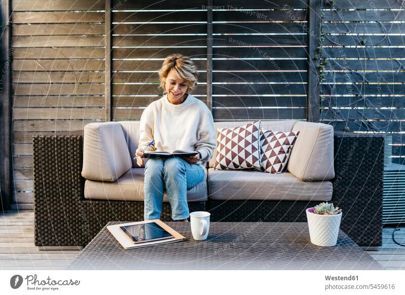 Lächelnde Frau schreibt Tagebuch, während sie auf dem Sofa im Dachgarten sitzt Farbaufnahme Farbe Farbfoto Farbphoto Außenaufnahme außen draußen im Freien
