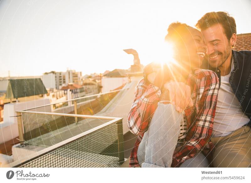 Glückliches, liebevolles junges Paar sitzt bei Sonnenuntergang auf dem Dach entspannen relaxen knuddeln schmusen sitzend Arm umlegen Umarmung Umarmungen abends