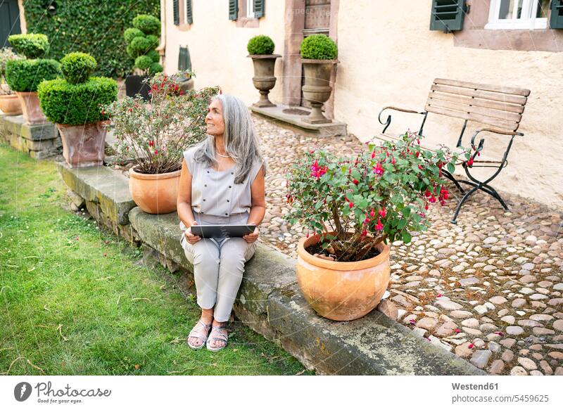 Frau mit langen grauen Haaren sitzt auf der Terrasse eines Landhauses und benutzt ein Tablet weiblich Frauen sitzen sitzend lächeln Landhäuser Landhaeuser