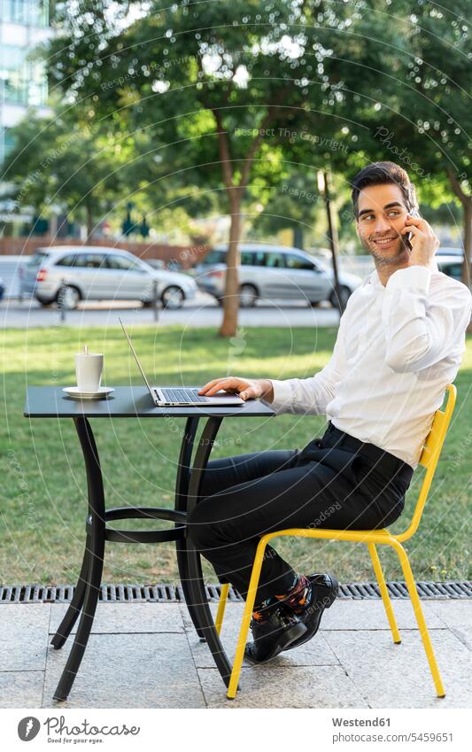 Gutaussehender Geschäftsmann mit Laptop auf dem Tisch, der im Außencafé über Handy telefoniert Farbaufnahme Farbe Farbfoto Farbphoto Spanien Außenaufnahme außen