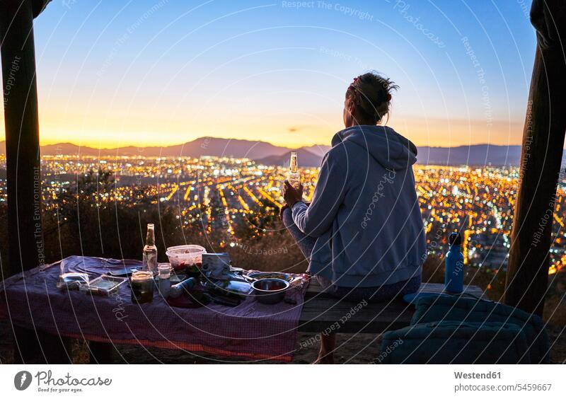 Chile, Santiago, Frau trinkt ein Bier in den Bergen über der Stadt bei Sonnenuntergang trinken staedtisch städtisch Sonnenuntergänge Außenaufnahme draußen