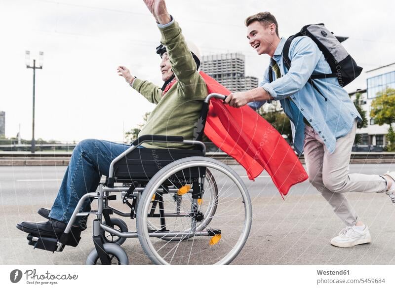 Junger Mann schiebt älteren Mann, der als Superheld verkleidet in einem Rollstuhl sitzt Leute Menschen People Person Personen Europäisch Kaukasier kaukasisch 2