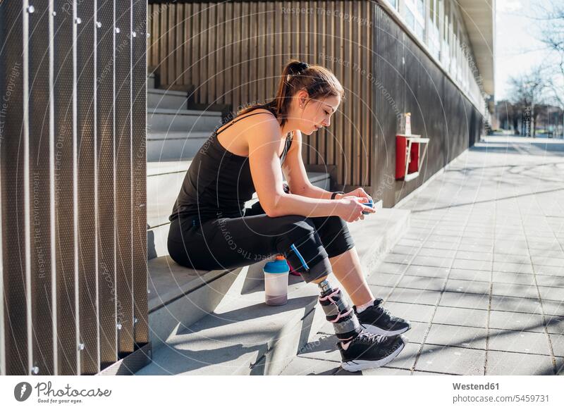 Sportliche junge Frau mit Beinprothese sitzt auf einer Treppe in der Stadt und benutzt ein Smartphone Telekommunikation telefonieren Handies Handys Mobiltelefon