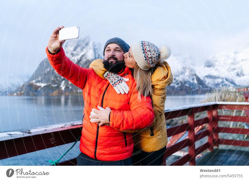 Touristenpaar macht ein Selfie in Hamnoy, Lofoten, Norwegen Jacken Telekommunikation telefonieren Handies Handys Mobiltelefon Mobiltelefone Arm umlegen Umarmung