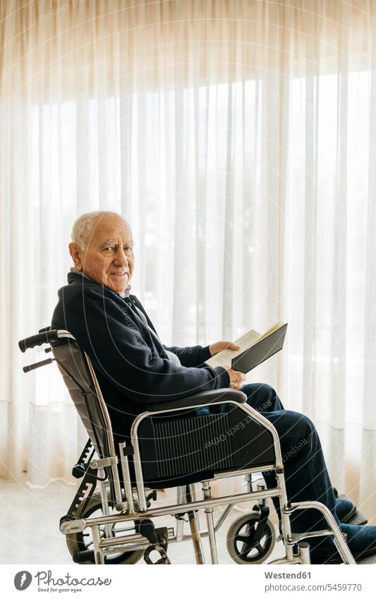 Porträt eines lächelnden älteren Mannes, der mit einem Buch im Rollstuhl sitzt Leute Menschen People Person Personen Europäisch Kaukasier kaukasisch 1 Ein