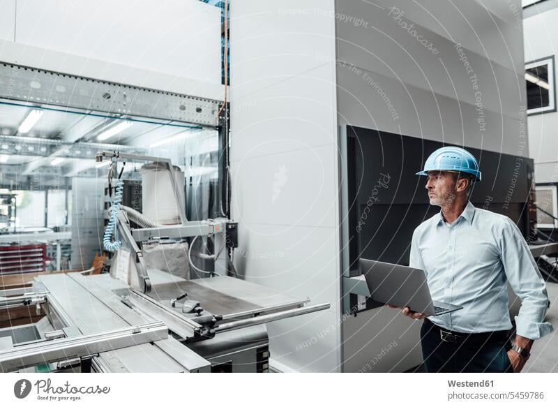 Männlicher Ingenieur mit Laptop beim Betrachten von Fertigungsmaschinen in der Industrie Farbaufnahme Farbe Farbfoto Farbphoto Deutschland Innenaufnahme