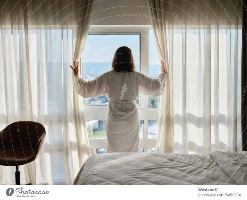 Frau öffnet weißen Vorhang, während sie durch das Fenster eines Luxushotelzimmers blickt Farbaufnahme Farbe Farbfoto Farbphoto Innenaufnahme Innenaufnahmen