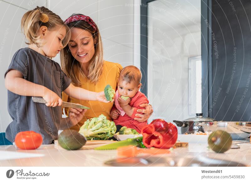 Mutter hält kleines Mädchen, während sie die Tochter beim Gemüseschneiden auf der Kücheninsel anleitet Farbaufnahme Farbe Farbfoto Farbphoto Innenaufnahme