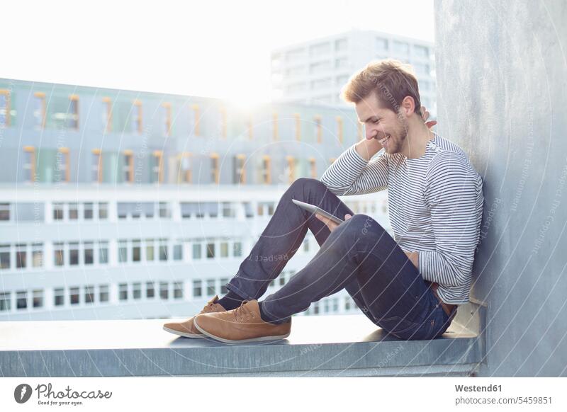 Glücklicher junger Mann sitzt an einer Wand und benutzt ein Tablet glücklich glücklich sein glücklichsein sitzen sitzend Mauer Mauern Tablet Computer Tablet-PC