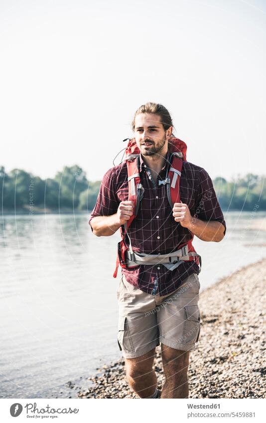 Junger Mann mit Rucksack beim Spaziergang am Flussufer Rucksäcke Fluesse Fluß Flüsse gehen gehend geht Männer männlich Gewässer Wasser Erwachsener erwachsen