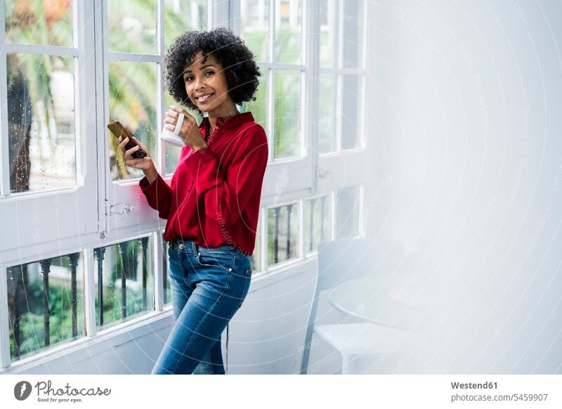 Porträt der lächelnden Frau mit Tasse Kaffee und Handy stehen am Fenster zu Hause stehend steht Kaffeetasse Kaffeetassen weiblich Frauen Zuhause daheim Portrait