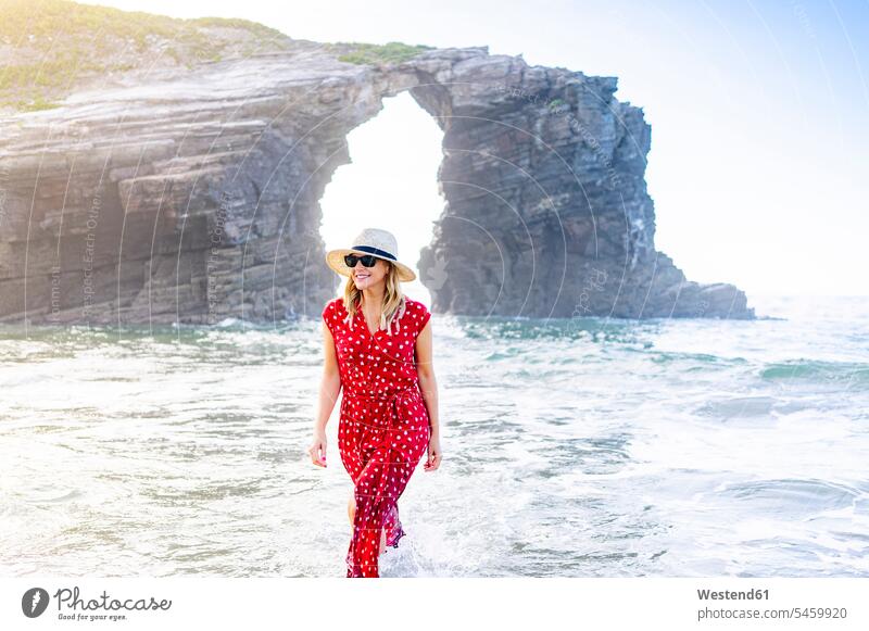 Blonde Frau in rotem Kleid und Hut, die am Strand spazieren geht, Natürlicher Bogen an der Playa de Las Catedrales, Spanien Brillen Sonnenbrillen freuen Glück