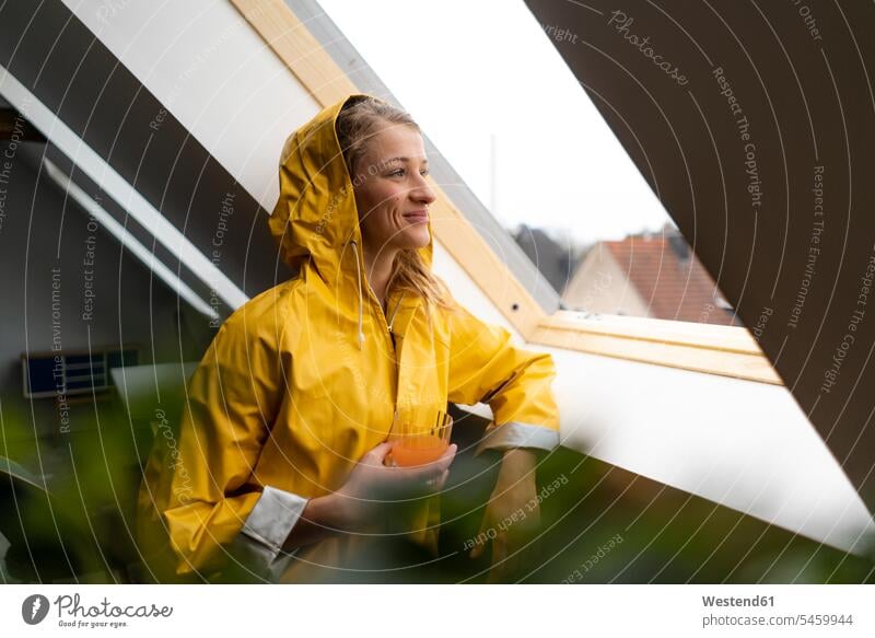 Lächelnde junge Frau im Regenmantel schaut aus dem Dachbodenfenster Single Alleinstehende Unverheirateter Singles Alleinstehender Regenjacke Regenjacken Wohnung