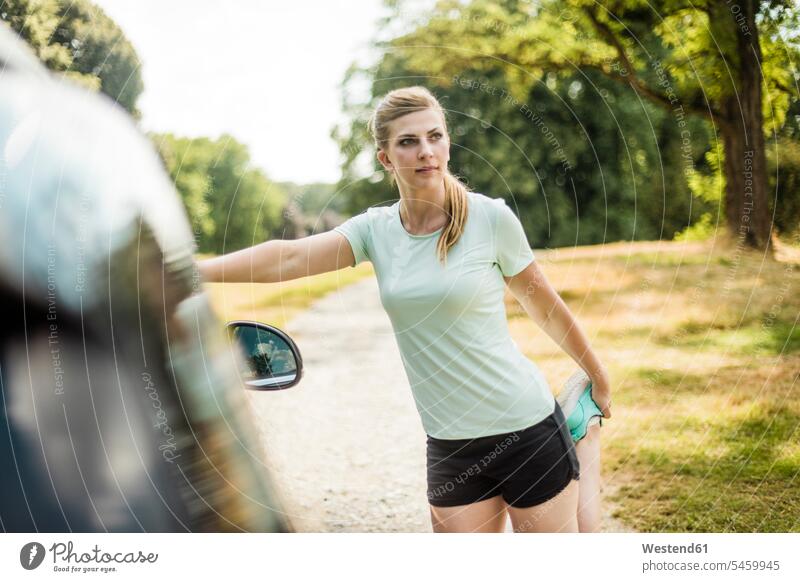 Sportliche junge Frau beim Dehnen an einem Auto in einem Park Wagen PKWs Automobil Autos sportlich weiblich Frauen dehnen strecken Parkanlagen Parks