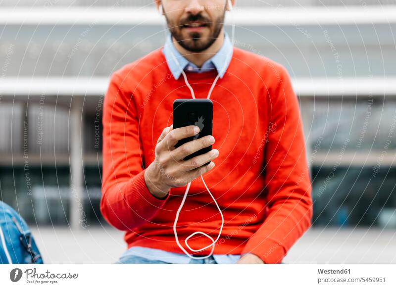 Lässiger Geschäftsmann, der auf einer Bank in der Stadt sitzt und sein Smartphone und Kopfhörer benutzt Urban städtisch Urbanität Urbanitaet Ohrhörer iPhone