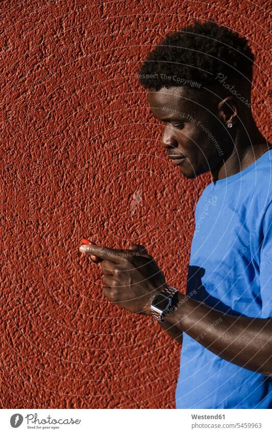 Nahaufnahme eines jungen Mannes, der bei Sonnenschein ein Smartphone gegen eine rote Wand in der Stadt benutzt Farbaufnahme Farbe Farbfoto Farbphoto Spanien