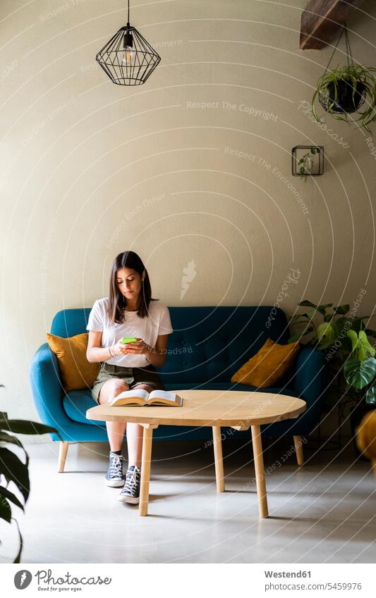 Junge Frau sitzt auf einer Couch und benutzt ein Mobiltelefon Informationen Bücher T-Shirts Couches Liege Sofas Telekommunikation telefonieren Handies Handys
