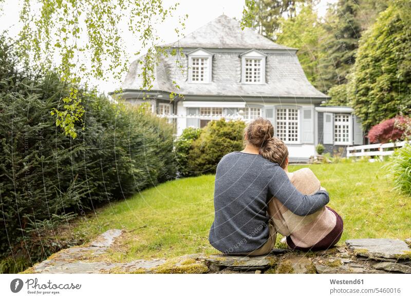 Rückansicht eines Paares, das auf einer Mauer im Garten sitzt sitzend Arm umlegen Umarmung Umarmungen entspannen relaxen entspanntheit relaxt geniessen Genuss