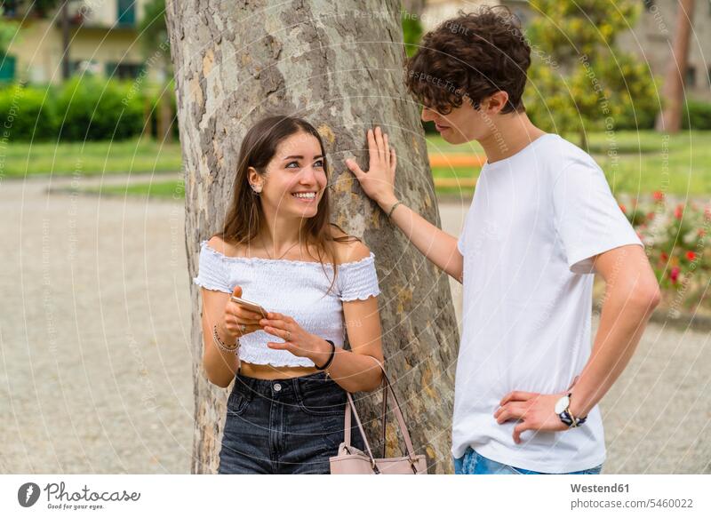 Junges Paar in einem Park, Frau zeigt ihm ihr Smartphone und lächelt Telekommunikation telefonieren Handies Handys Mobiltelefon Mobiltelefone sommerlich