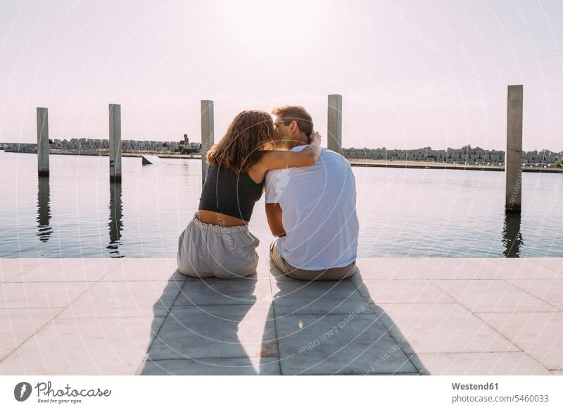 Rückansicht eines jungen Paares, das auf einer Mole am Meer sitzt und sich küsst Touristen Kuss Küsse sitzend Arm umlegen Umarmung Umarmungen Jahreszeiten