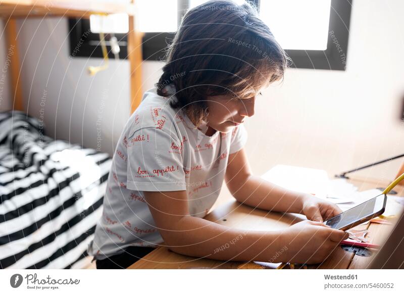 Junge sitzt zu Hause am Schreibtisch und benutzt ein Smartphone Informationen T-Shirts Tische Arbeitstisch Schreibtische Telekommunikation Handies Handys