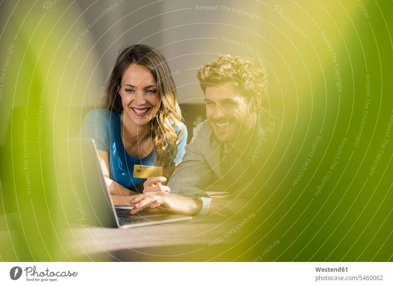 Lächelndes Paar beim Online-Shopping mit Laptop auf dem Sofa zu Hause Notebook Laptops Notebooks liegen liegend liegt Zuhause daheim Pärchen Paare Partnerschaft