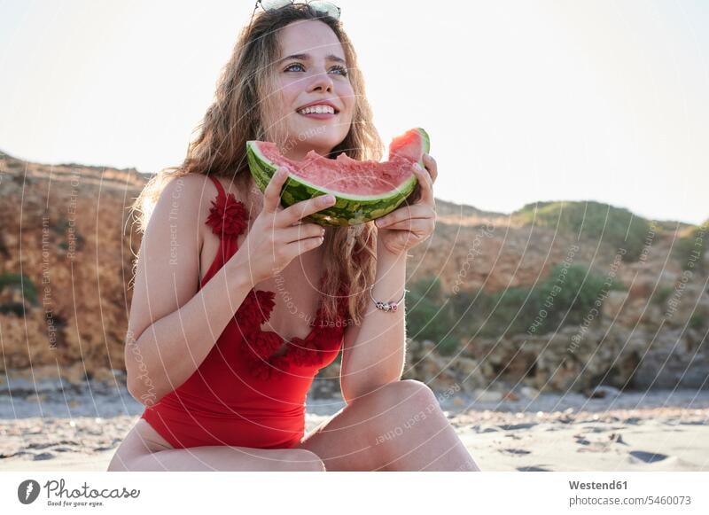 Glückliche junge Frau hält Wassermelonenscheibe am Strand Badebekleidung Badeanzuege Badeanzüge sitzend sitzt sommerlich Sommerzeit entspannen relaxen