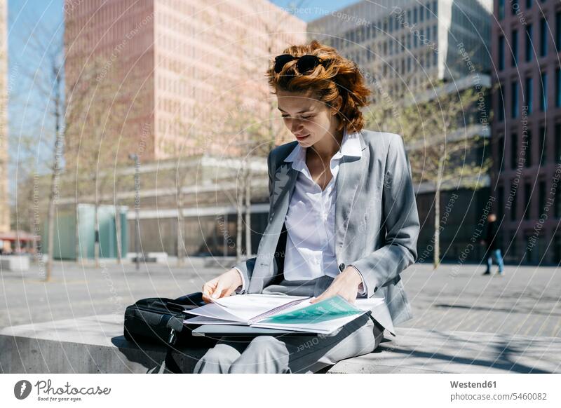 Junge Geschäftsfrau liest Dokumente, sitzt auf einer Bank in der Stadt an einem sonnigen Tag Hosenanzug Urban städtisch Urbanität Urbanitaet sitzen sitzend