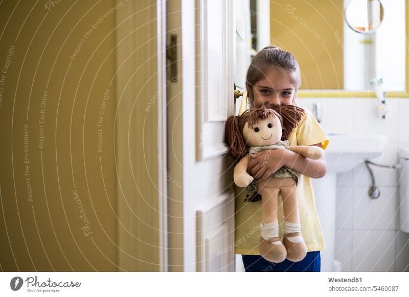 Porträt eines lächelnden kleinen Mädchens, das zu Hause im Türrahmen steht und eine Puppe hält Zuhause daheim weiblich stehen stehend Portrait Porträts