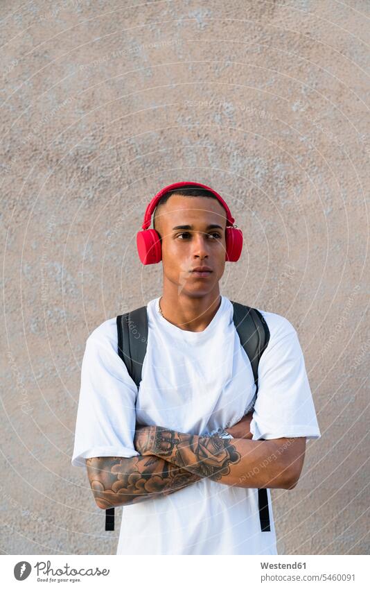 Porträt eines tätowierten jungen Mannes, der mit drahtlosen roten Kopfhörern Musik hört Leute Menschen People Person Personen gemischtrassig 1 Ein ein Mensch