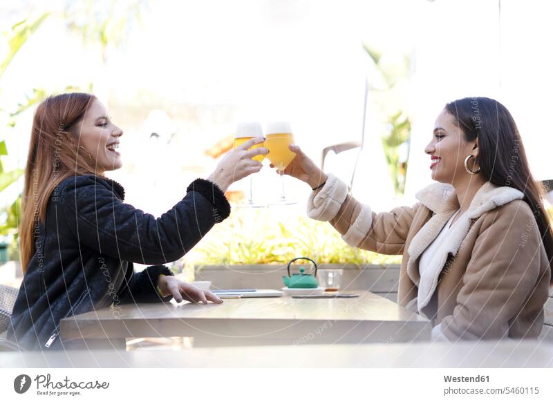 Zwei Freundinnen, die draußen in einem Café mit Biergläsern klimpern Freunde Kameradschaft Gläser Trinkglas Trinkgläser Jacken Tische sitzend sitzt entspannen