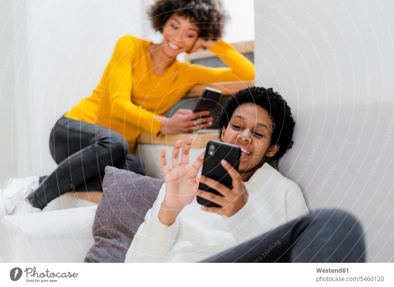 Zwei junge Frauen entspannen sich zu Hause mit ihren Smartphones Freunde Kameradschaft Freundin Couch Couches Liege Sofas Telekommunikation Handies Handys