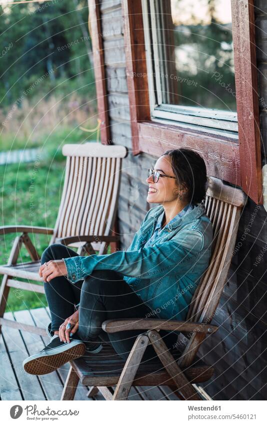 Glückliche junge Frau sitzt auf der Veranda eines Holzhauses junge Frauen glücklich glücklich sein glücklichsein Holzhäuser Holzhaeuser offenes Lächeln lachen