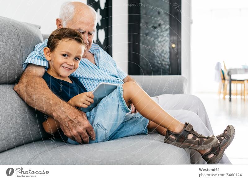 Porträt des zufriedenen kleinen Jungen mit digitaler Tablette sitzt neben seinem Großvater auf der Couch zu Hause Opa Großpapa Großpapas Opas Opi Großväter Opis