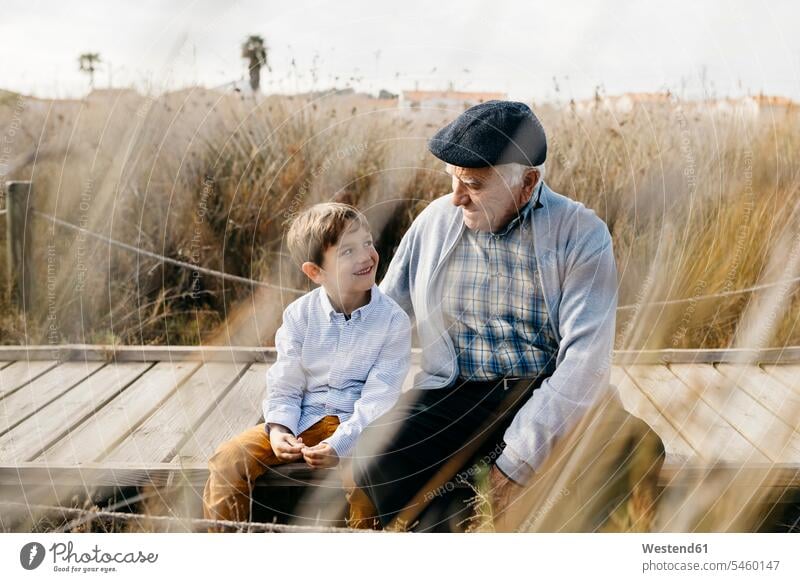Großvater sitzt mit seinem Enkel auf der Strandpromenade und spricht Kommunikation Gras Gräser Graeser Nähe nah innig Betreuung betreuen Fürsorge kümmern