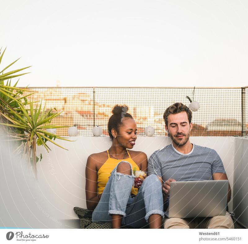 Junges Paar mit Laptop und Kopfhörern sitzt abends auf dem Dach Polster Rechner Laptops Notebook Notebooks Medium Videofilm Videos entspannen relaxen hoeren