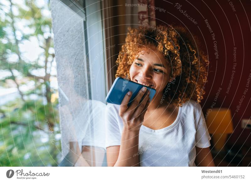 Junge Frau mit lockigem Haar sendet Sprachnachricht mit Handy neben dem Fenster Sprachnachrichten voice mails Voicemail Voicemails Locken lockiges Haar