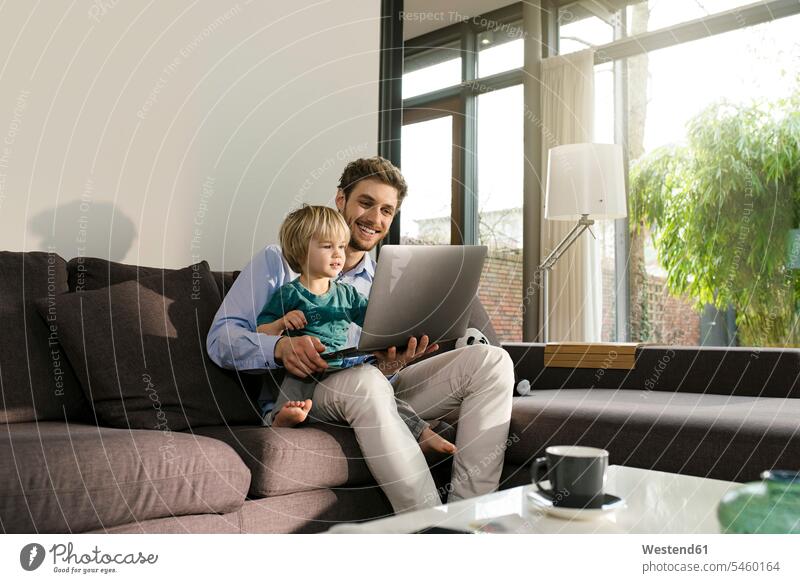 Vater und Sohn schauen sich zu Hause auf der Couch einen Laptop an Söhne Zuhause daheim sitzen sitzend sitzt Notebook Laptops Notebooks ansehen Sofa Couches