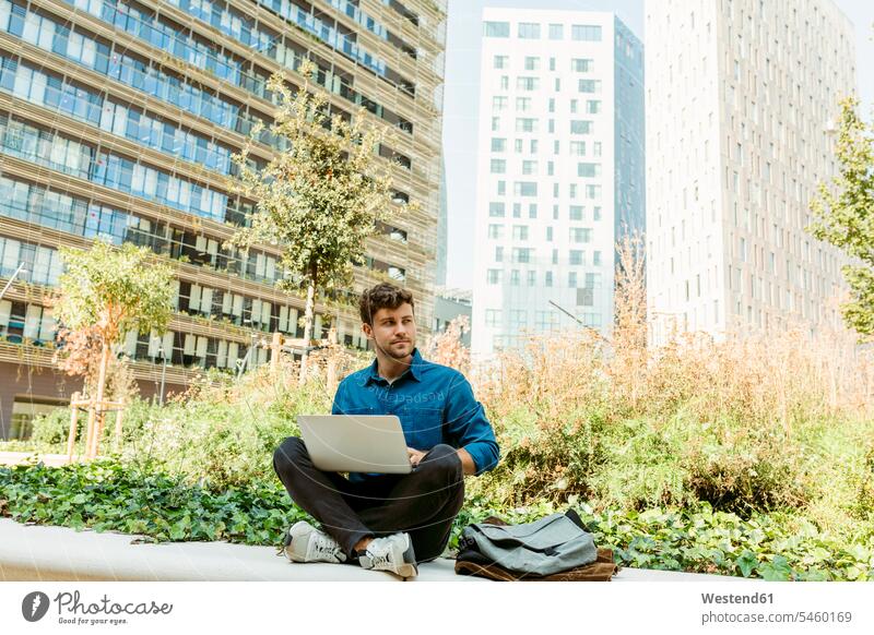 Geschäftsmann schaut weg, während er mit seinem Laptop an der Stützmauer vor dem Bürogebäude sitzt Farbaufnahme Farbe Farbfoto Farbphoto Spanien Geschäftsmänner