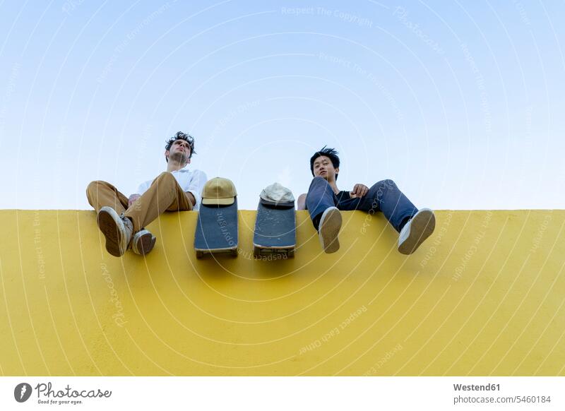 Zwei junge Männer mit Skateboards sitzen auf einer hohen gelben Wand Freunde Rollbretter ausruhen Rast Erholung erholen Mauer Mauern Mann männlich hoch sitzend