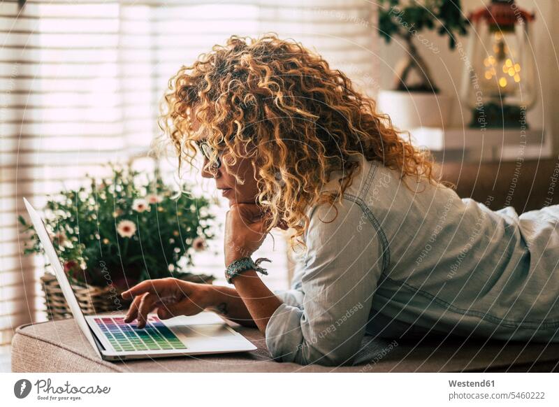 Lächelnde Frau liegt auf der Couch zu Hause mit Laptop Fenster Mobilität mobil Locken lockiges Haar gelockte Haare gelocktes Haar Verbindung verbunden verbinden