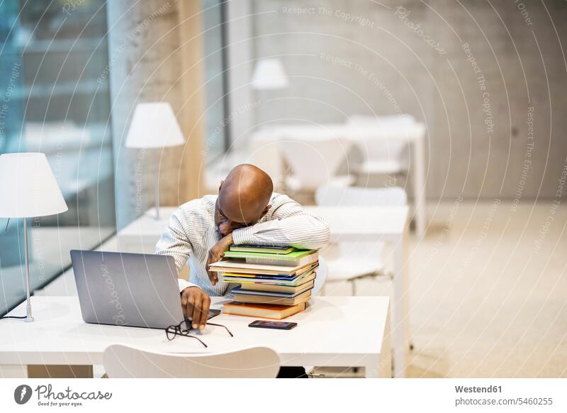 Erschöpfter reifer Mann sitzt am Schreibtisch und stützt sich auf einen Stapel von Büchern Erschöpfung erschöpft sitzen sitzend aufstützen aufgestuetzt