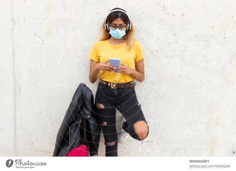 Junge Frau trägt Schutzmaske, während sie mit ihrem Gepäck mit einem Smartphone an der Bushaltestelle gegen die weiße Wand telefoniert Farbaufnahme Farbe