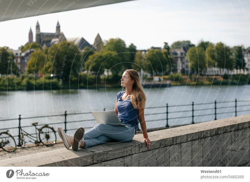 Niederlande, Maastricht, junge Frau sitzt mit Laptop auf einer Mauer am Flussufer weiblich Frauen Notebook Laptops Notebooks Fluesse Fluß Flüsse sitzen sitzend
