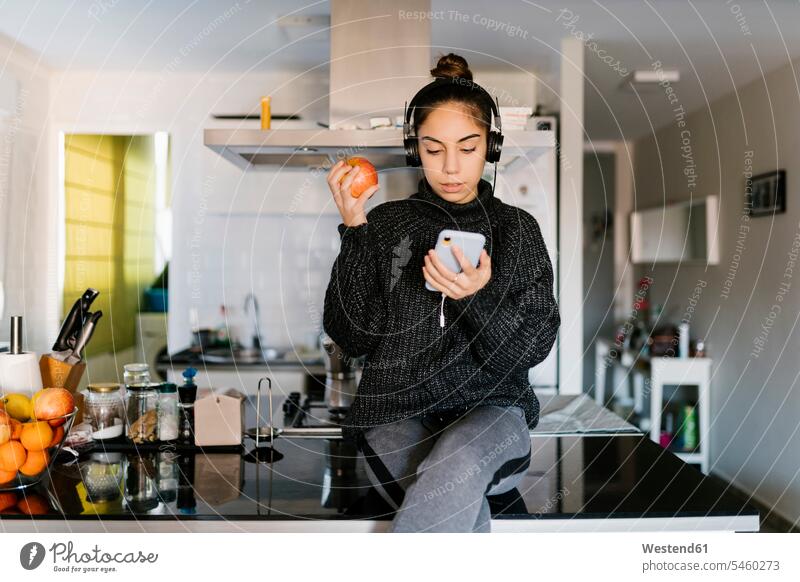 Teenagerin mit Kopfhörer, die einen Apfel hält, während sie zu Hause ein Mobiltelefon benutzt Farbaufnahme Farbe Farbfoto Farbphoto Innenaufnahme Innenaufnahmen