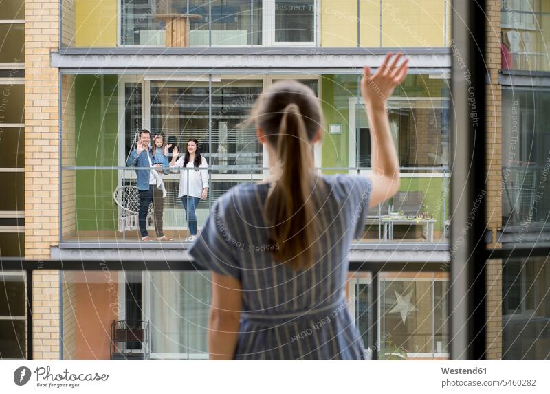 Rückenansicht einer Frau, die auf dem Balkon steht und ihren Nachbarn zuwinkt stehend Muße Lifestyles außen draußen im Freien staedtisch städtisch Urbanität