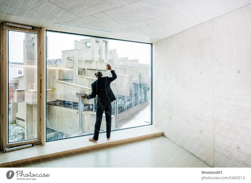 Rückenansicht eines barfuß gehenden Geschäftsmannes mit Virtual-Reality-Brille, der auf dem Fensterbrett steht und aus dem Fenster schaut Businessmann
