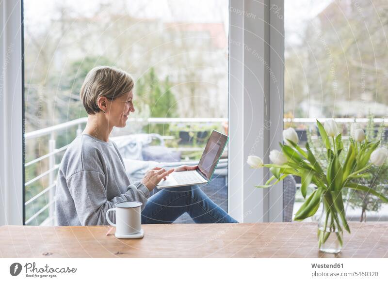 Reife Frau, die von zu Hause aus arbeitet, einen Laptop benutzt und auf einer Bank sitzt Job Berufe Berufstätigkeit Beschäftigung Jobs Tische Holztische Rechner