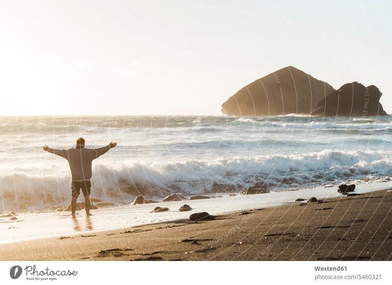 Mann steht bei Sonnenuntergang mit ausgestreckten Armen am Strand, Sao-Miguel-Insel, Azoren, Portugal Touristen entspannen relaxen abends entspanntheit relaxt